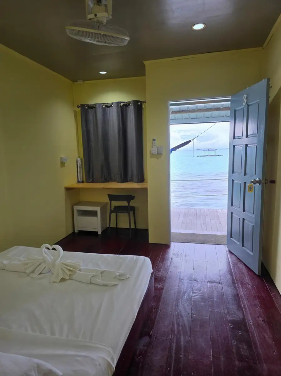 A cozy double room at Bigfin Water Bungalow over the Celebes Sea offering a large bed, traditional style wooden desk, hardwood floors, AC, and an open door opening to the overwater deck.