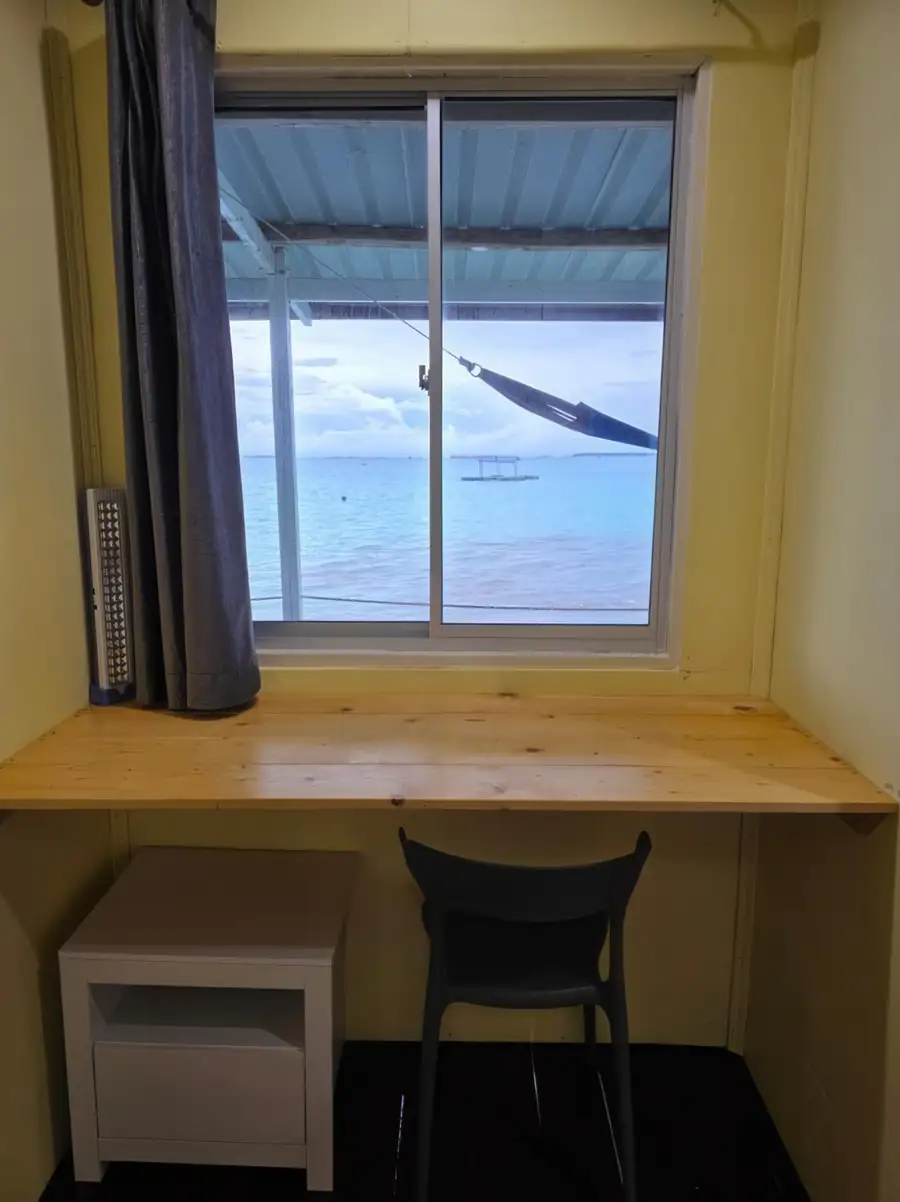 A practical bedroom workspace at Bigfin Water Bungalow near Tun Sakaran Marine Park featuring a modern style wooden desk, solid wood floors, AC, and a sliding window with stunning seaviews.