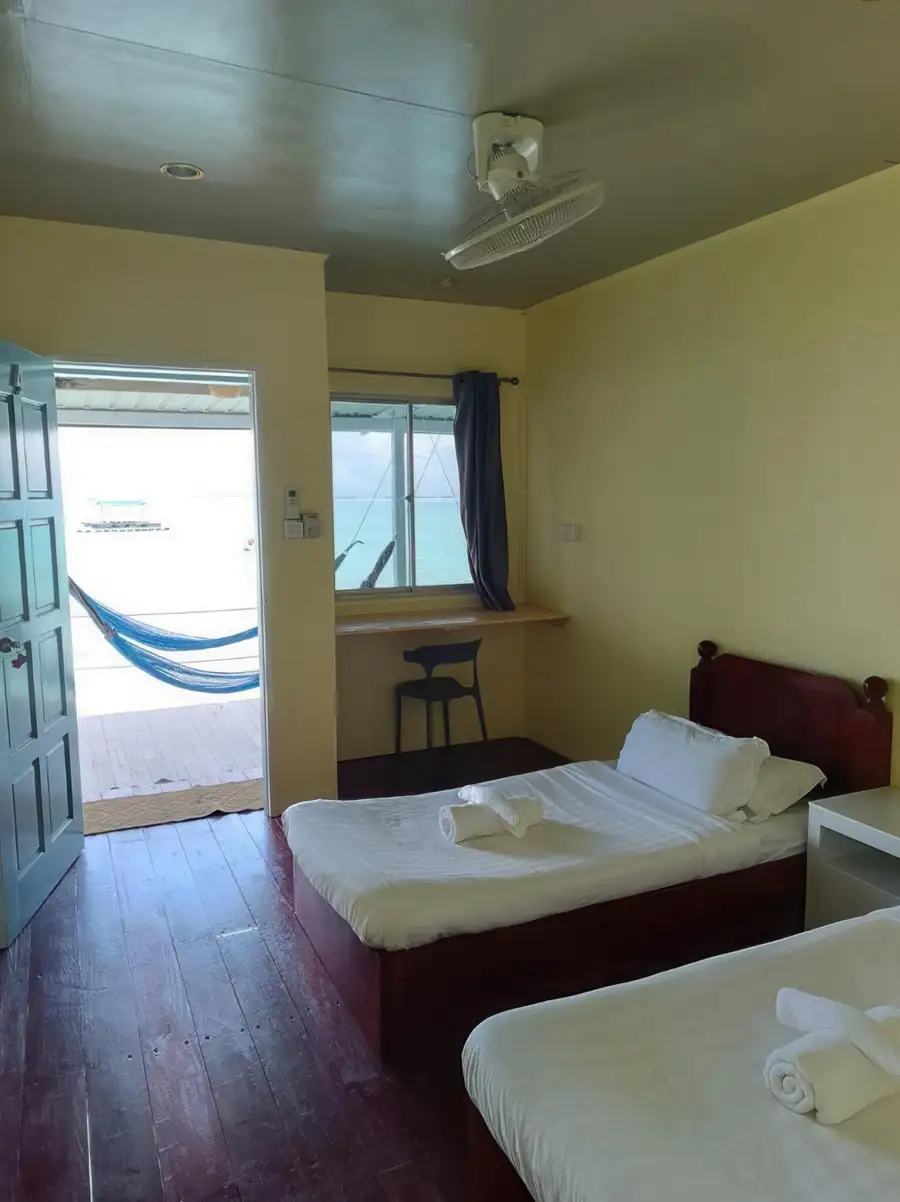 A relaxing bedroom at Bigfin Water Bungalow near Pulau Bum Bum showcasing two beds, a traditional style wooden desk, dark hardwood floors, air con, and a door opening to the deck.
