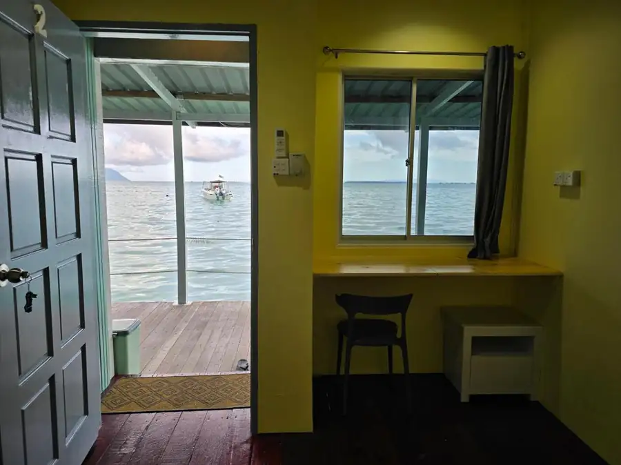 A cozy room entryway at Bigfin Water Bungalow over the Celebes Sea featuring a wooden desk, dark timber floors, AC, and an open wooden door leading to the overwater deck.